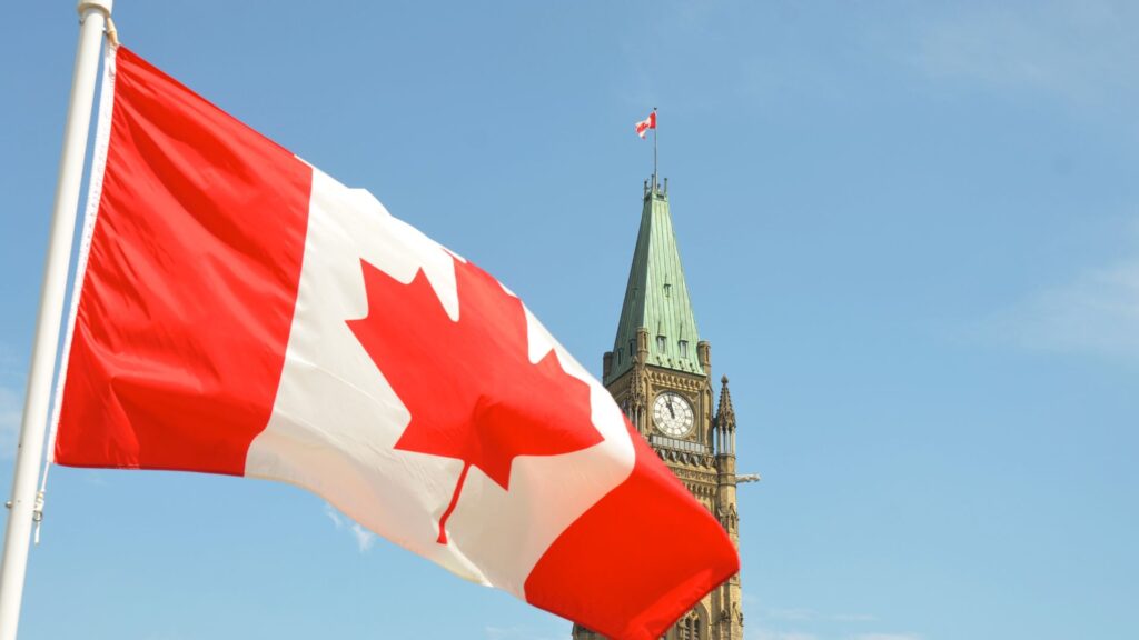 The Canadian national flag, with its distinctive red and white colors and maple leaf emblem, is fluttering on a flagpole in the foreground. Behind it, the iconic Peace Tower of Parliament Hill in Ottawa is visible against a clear blue sky