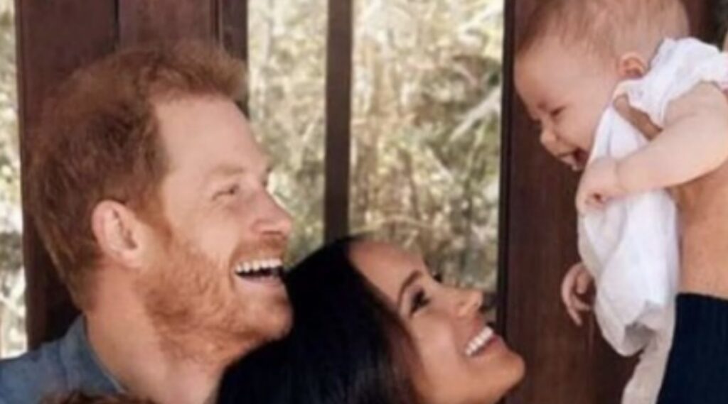 Prince Harry, Meghan Markle, and Princess Lilibet. Meghan is holding Lilibet up in the air and all three of them are smiling and laughing.