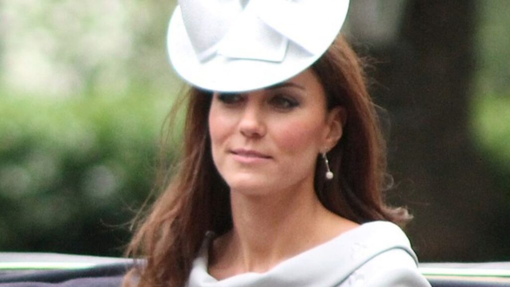 Kate Middleton in a horse and carriage. She is wearing a light blue fascinator and dress with pearl earrings.