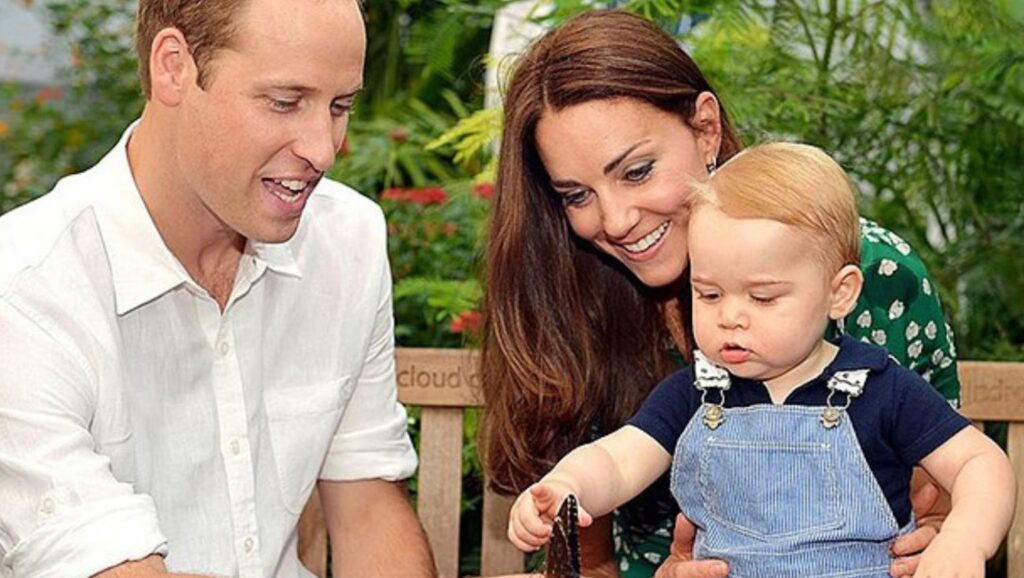 Prince William, Kate Middleton, and Prince George. Prince William has a butterfly on his hand and Prince George is sitting on Kate Middleton’s lap, pointing at the butterfly. They are all sat on a bench and there is lots of greenery behind them.