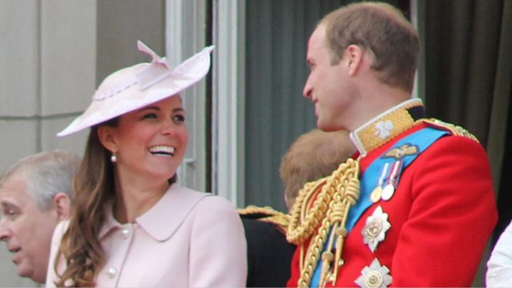 Kate Middleton and Prince William on the Buckingham Palace balcony. Kate is wearing a pink hat and coat and William is wearing a red military suit, with a blue sash, gold detailing, and medals.