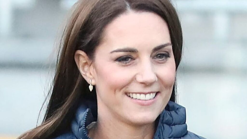 Kate Middleton smiling. She is wearing a blue puffer jacket and has heart earrings.