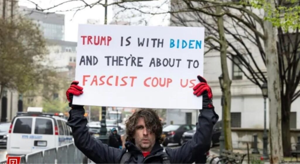 Max Azzarello during a protest. He is holding up a sign that says, “Trump is with Biden and they’re about to fascist coup us.”