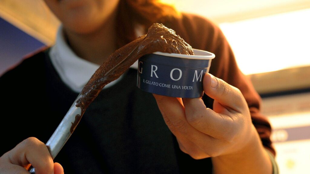 person is putting chocolate ice cream into a blue cardboard tub. She is using a silver spoon to do so.