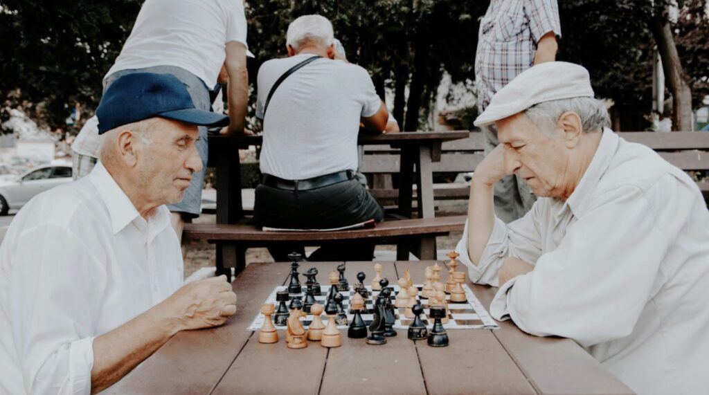 Two old men in hats play a game of chess opposite each other outside, with other men behind them.