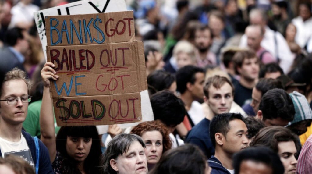 A dense crowd of protesters, with various expressions of concern and frustration, where one individual is holding up a cardboard sign that reads 'BANKS GOT BAILED OUT WE GOT SOLD OUT' in a public demonstration