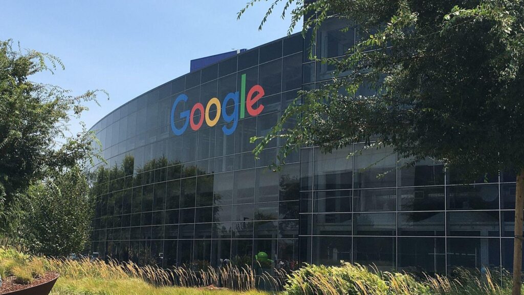 A photo showing the exterior of the Google headquarters building. The building has a curved glass facade with the colorful Google logo prominently displayed