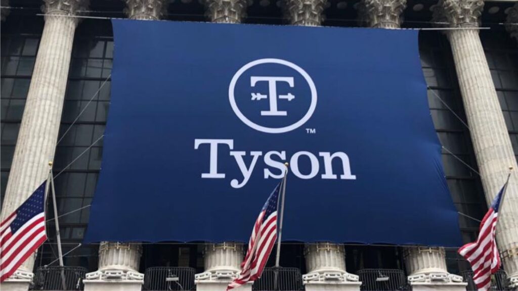 A large blue banner with the Tyson Foods logo is draped on the facade of a building with classical columns. Two American flags are flying on flagpoles in front of the building, on either side of the banner