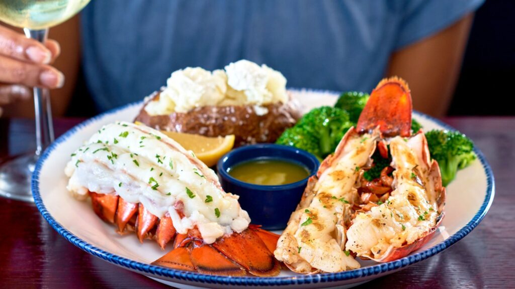 A plate with a half lobster and a steak garnished with chives, accompanied by broccoli, mashed potatoes, and a lemon wedge, served on a blue rimmed plate with a glass of white wine in the background