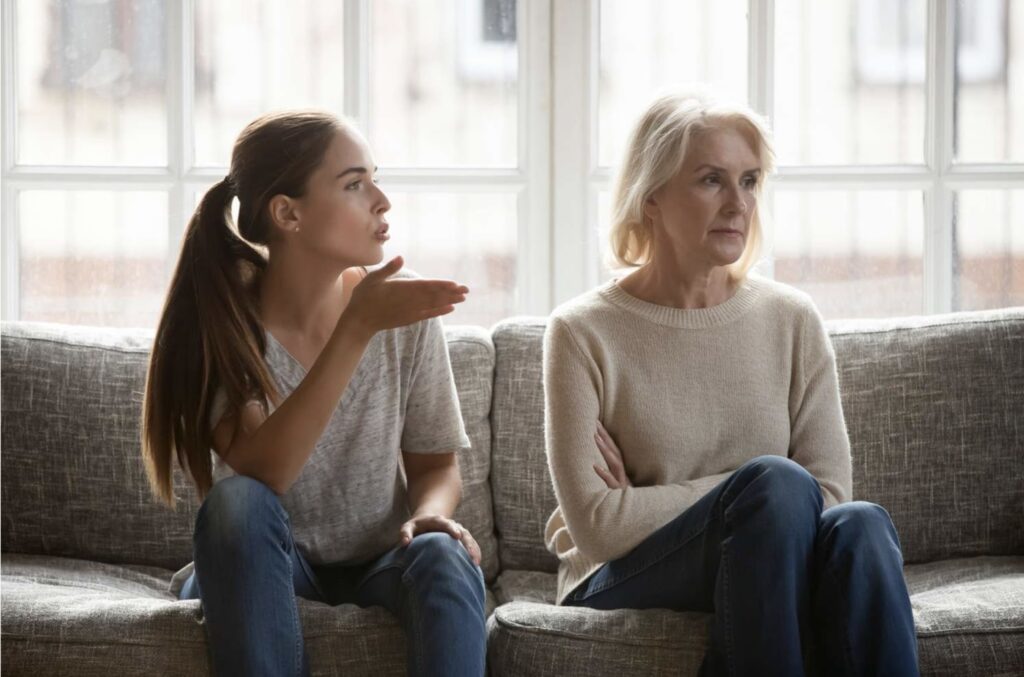 An adult woman sits on the couch with her mother, clearly expressing frustration as the mother looks disappointed