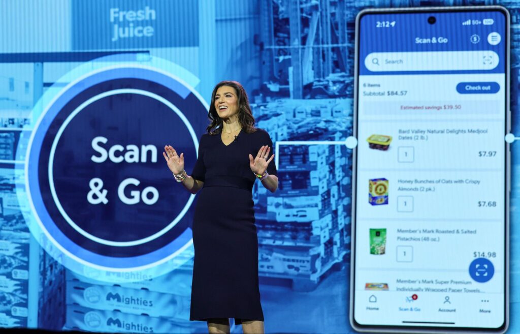 A female representative of Sam’s Club giving a presentation of the retailer’s Scan & Go app with a screen behind her showing images the store and a mobile app