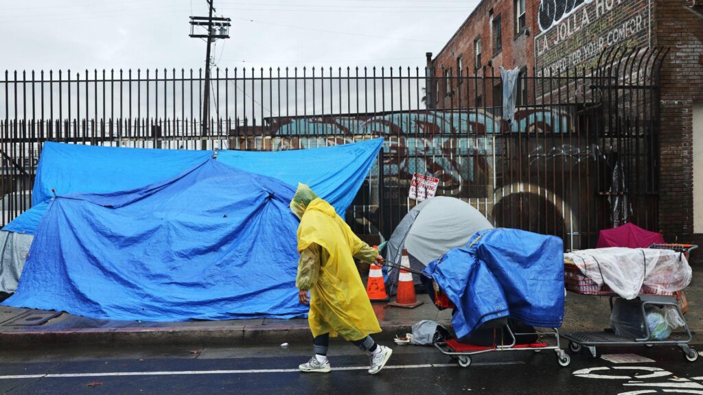 A person in a yellow anorak dragging a trolley down a street. They are walking past tents that have been set up on the side of the street.