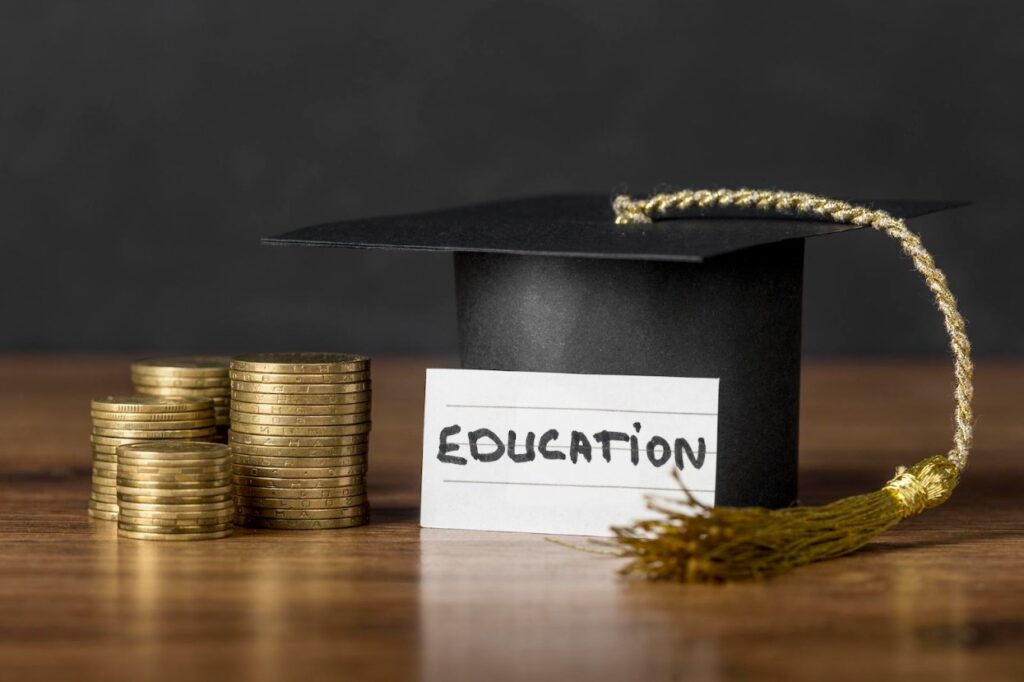 A black academic cap model next to a stack of gold coins with a scrap of paper with the writing Education in front of it