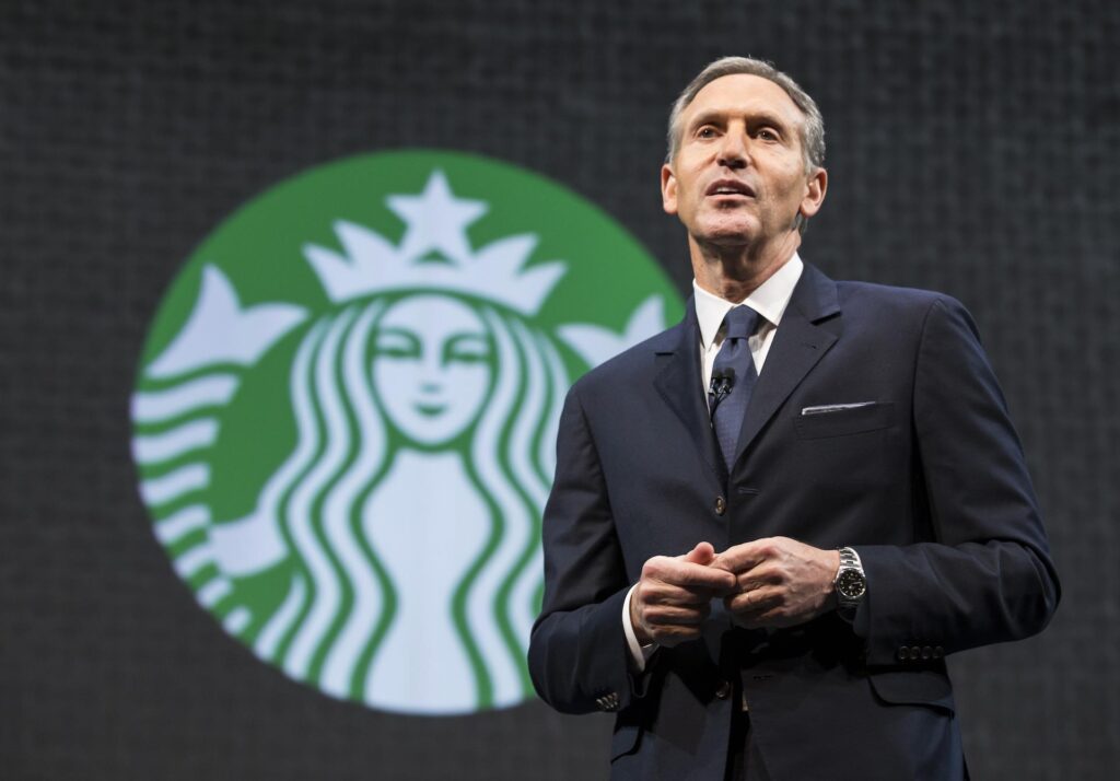 Howard Schultz wearing a suit and a tie standing in front of the green Starbucks logo