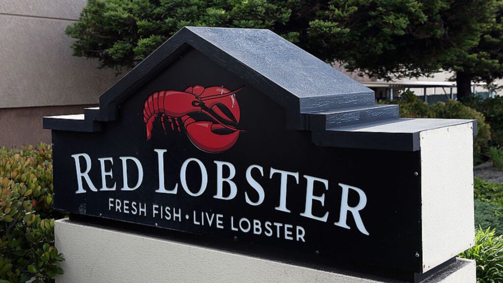 Close-up of a Red Lobster restaurant sign with the logo and text "Fresh Fish - Live Lobster" on a black background, set against a backdrop of shrubbery and a partially visible building