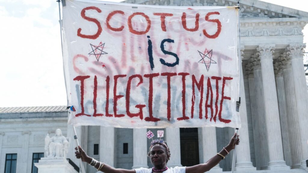 Protestor stood in front of the Supreme Court building with a large banner reading "SCOTUS is Illegitimate".