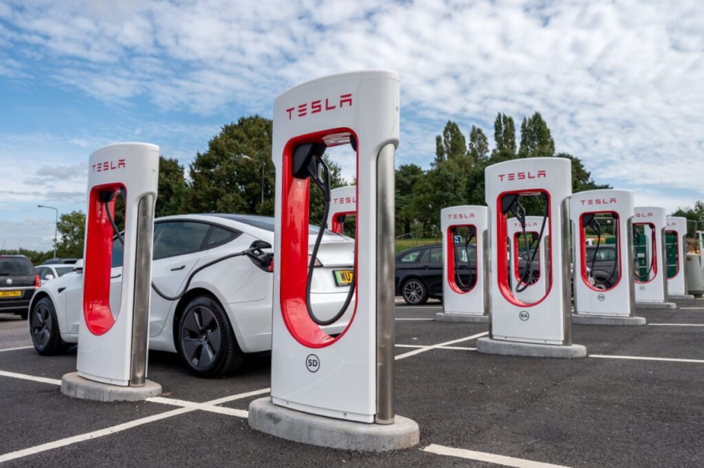 A row of white Tesla charging stations in a grey parking lot.