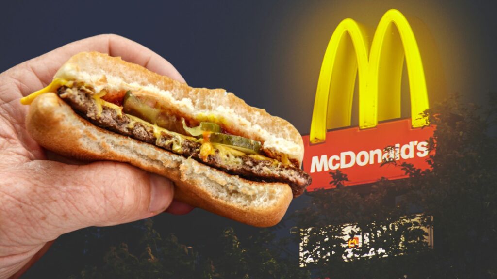 Close-up of a hand holding a partially eaten McDonald's burger with visible layers of beef patty, cheese, pickles, and condiments, with the McDonald's logo illuminated in the background at night