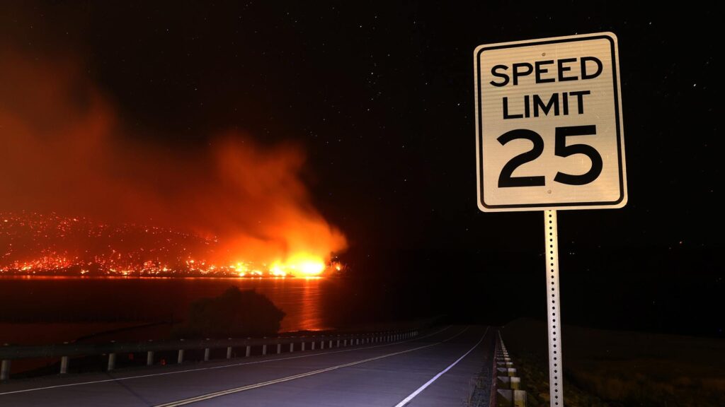 A fire burns in the distance as the viewer passes a speed limit sign on the road.