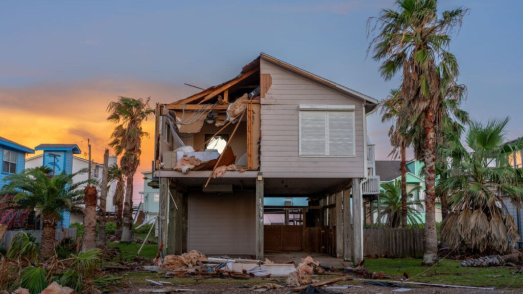 A house with severe structural damage including exposed interior rooms and missing walls, following a hurricane. Palm trees and neighboring houses in the background show signs of storm damage under a sunset sky