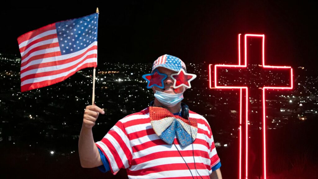 A person at a night parade wearing a striped red, white, and blue outfit with star-shaped glasses, and a mask, holding an American flag next to a large neon cross