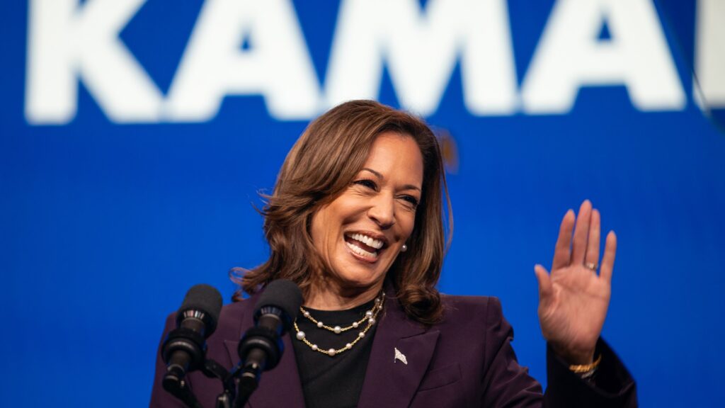 VP Kamala Harris waves on stage with her name behind her.