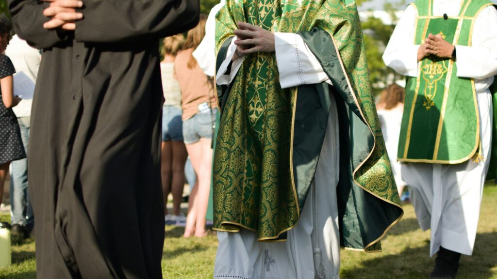 Priests in green vestments participating in an outdoor Catholic Mass with congregation members in the background