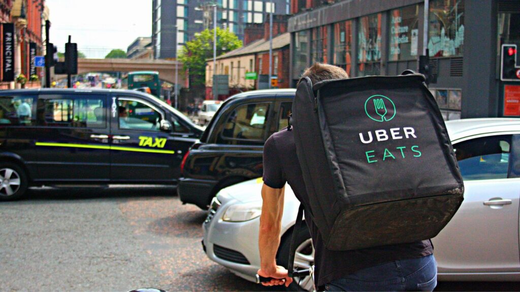 An Uber Eats delivery person walking on a city street with a large insulated backpack, navigating through traffic with cars and taxis around