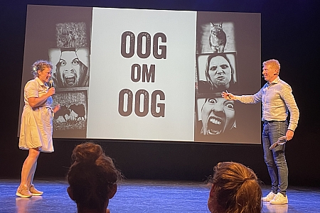 Op scherm staat: 'Oog om oog.' 
Frederik Imbo in gesprek met een vrijwilliger uit het publiek.