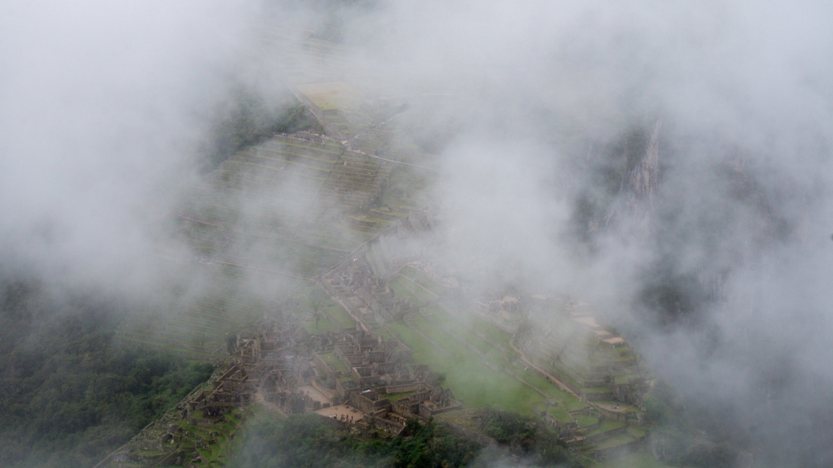 A close up of Machu Picchu taken from the top of Huayna Picchu on a misty day. The clouds mostly cover the ruins.
