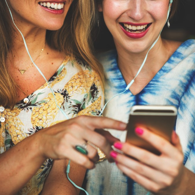 Two female friends sharing earbuds smiling and listening to a podcast