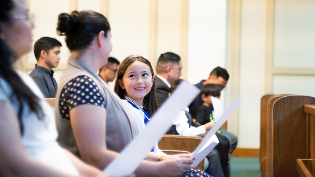 A worship service setting with a handful of people sitting on pews and a child smiling and looking at the woman next to her.