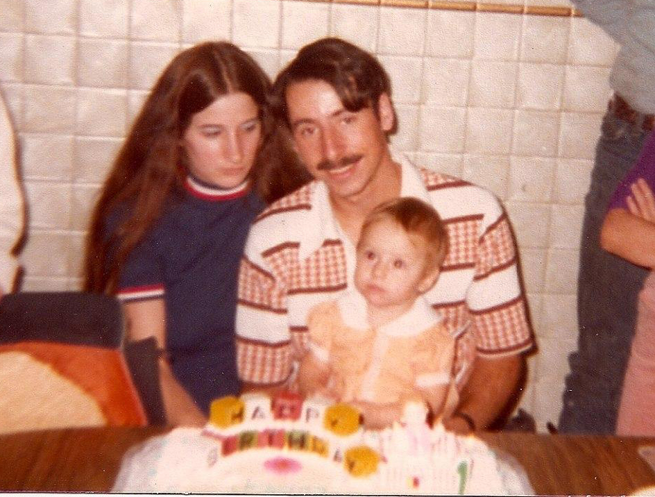 An old family photo with a mom, dad and child sitting in front of a birthday cake.