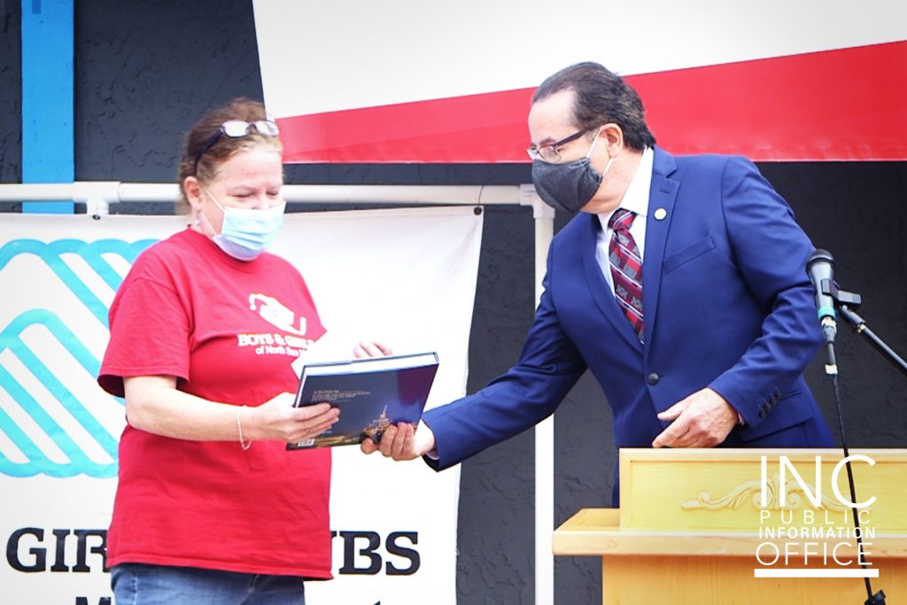 A man in a suit hands a woman a large book, both wearing masks standing behind a podium 