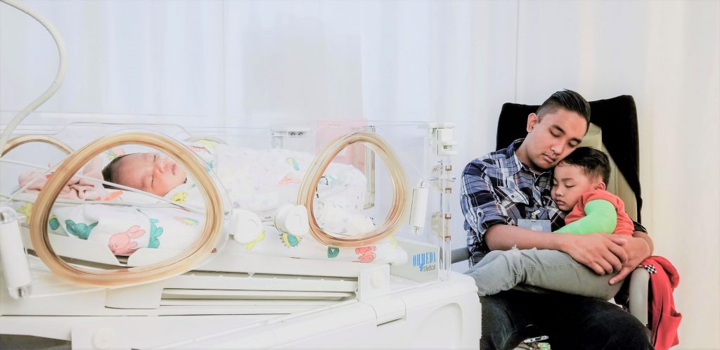 A baby in NICU while dad and son sleep next to bed.