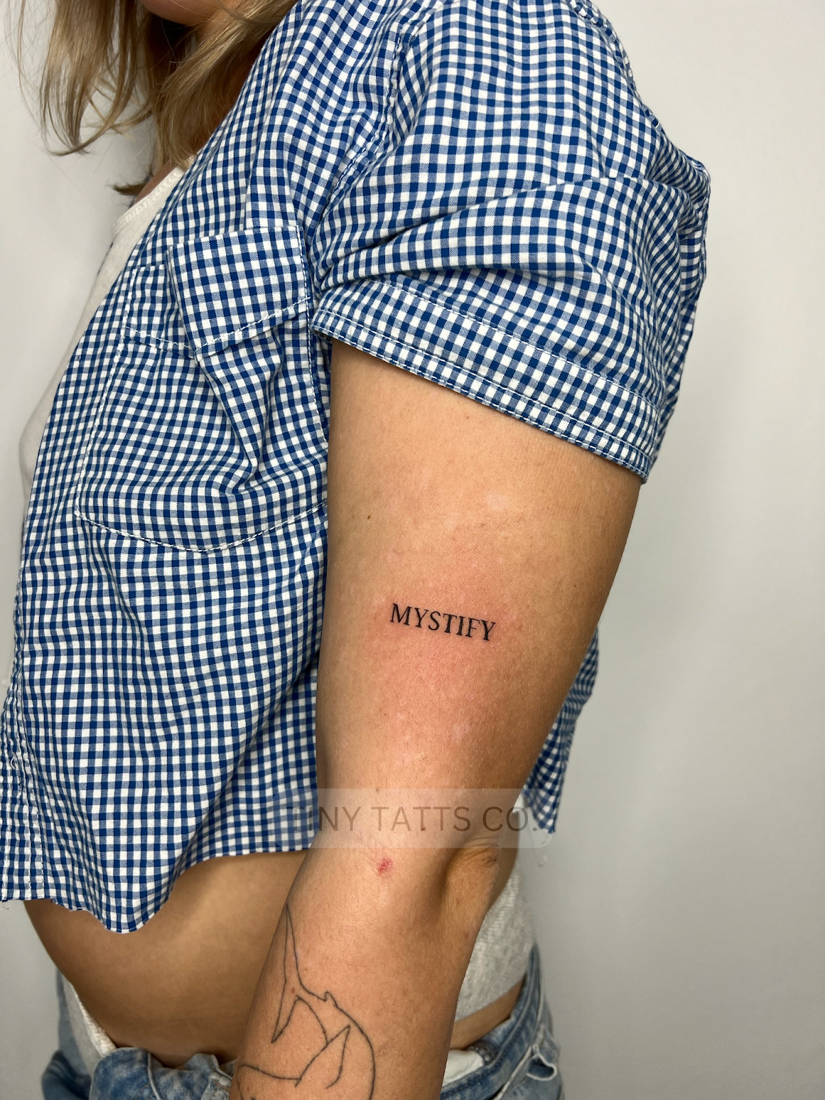 A close-up of a minimalist linework tattoo featuring the word 'Mystify' on the forearm.