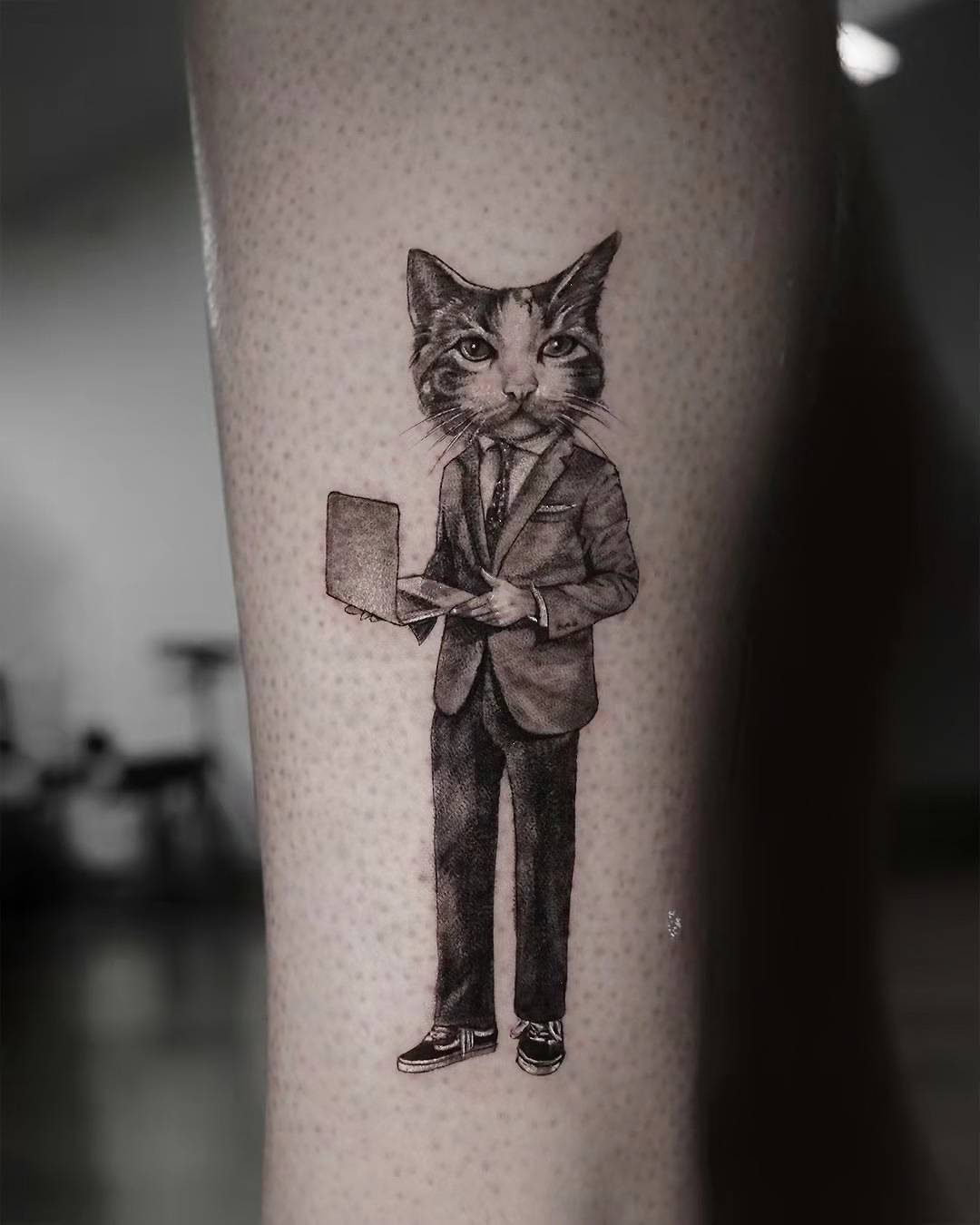 A detailed black and grey tattoo of a cat dressed in business attire holding a laptop.