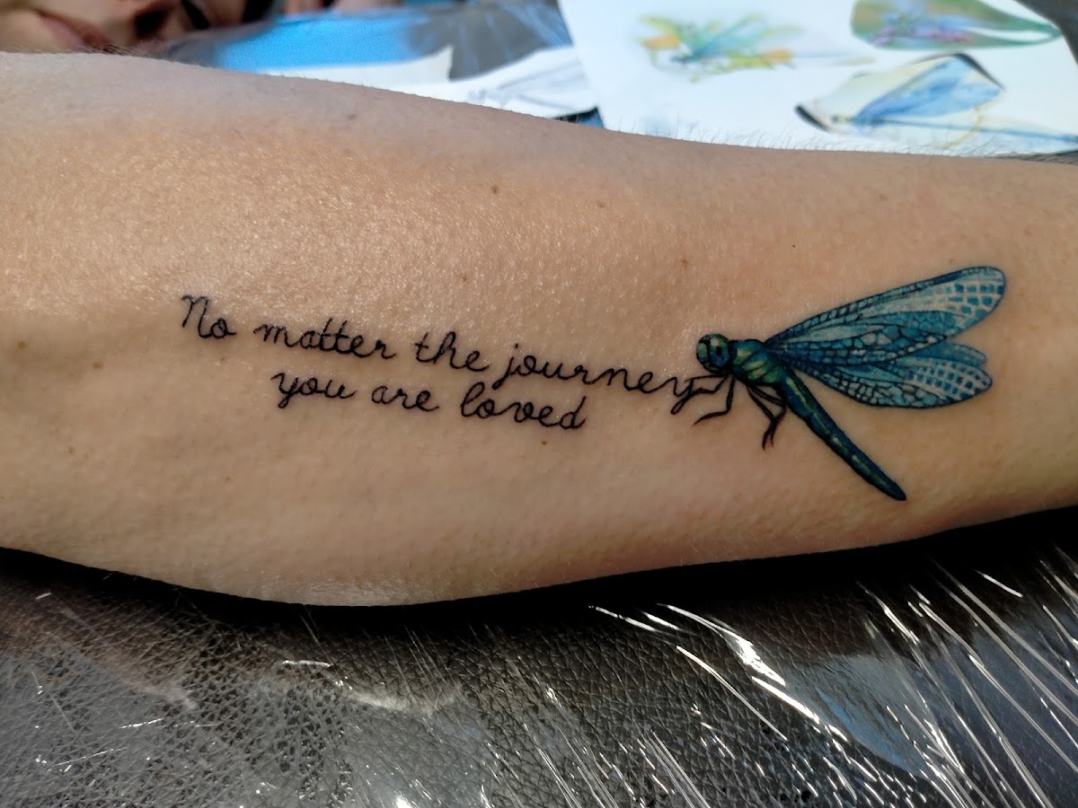 A delicate dragonfly tattoo with a motivational quote is inked on the forearm.