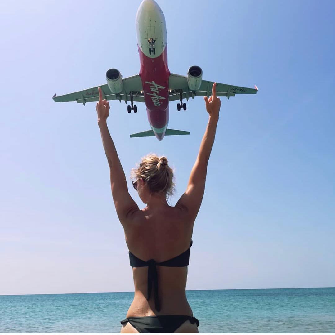 A female traveler posing with Air Asia aircraft crossing the sky at Mai Khao Beach