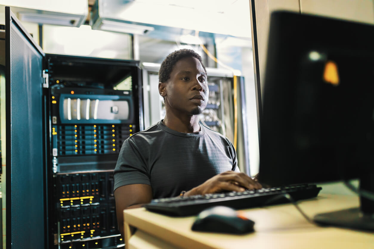 A man works on a computer with a large switchboard behind him. 