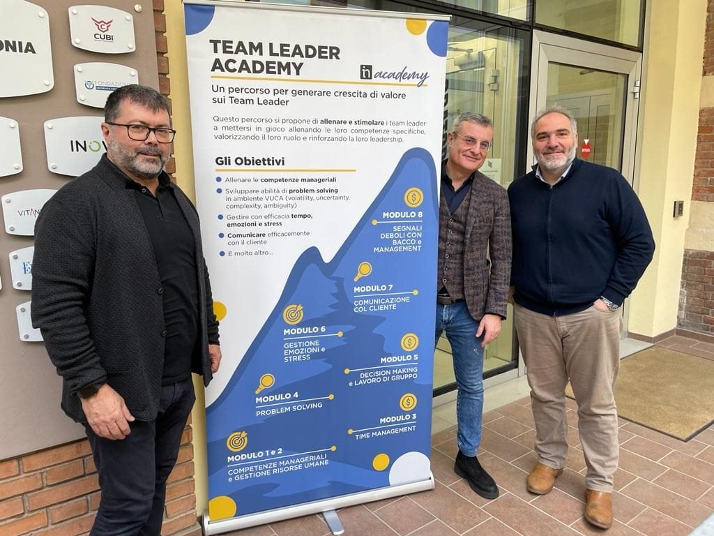 Tre uomini posano davanti a un banner di "Team Leader Academy".