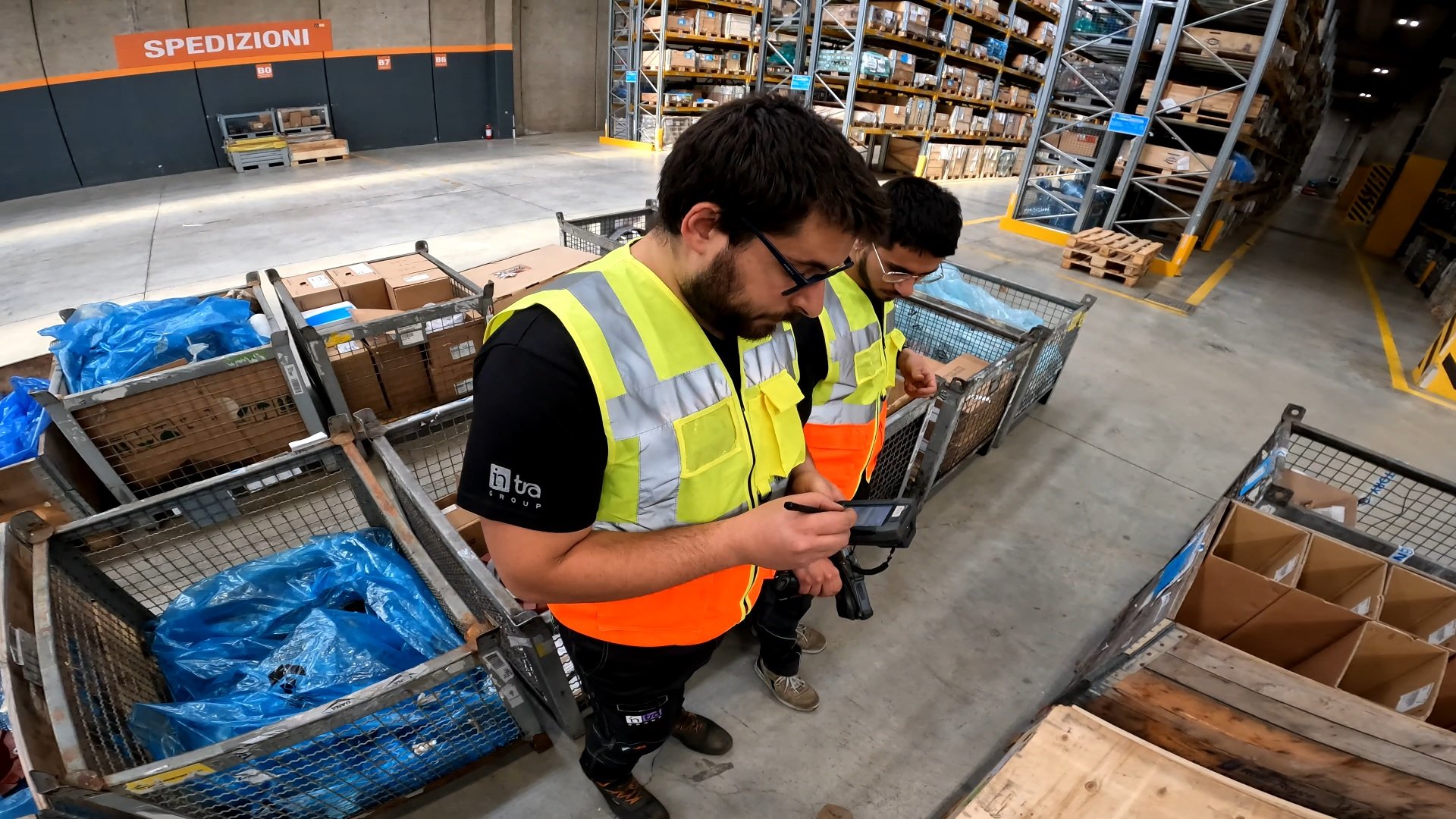 Due addetti al magazzino controllano la merce con un dispositivo elettronico. Indossano gilet ad alta visibilità. Sullo sfondo scaffali pieni.
