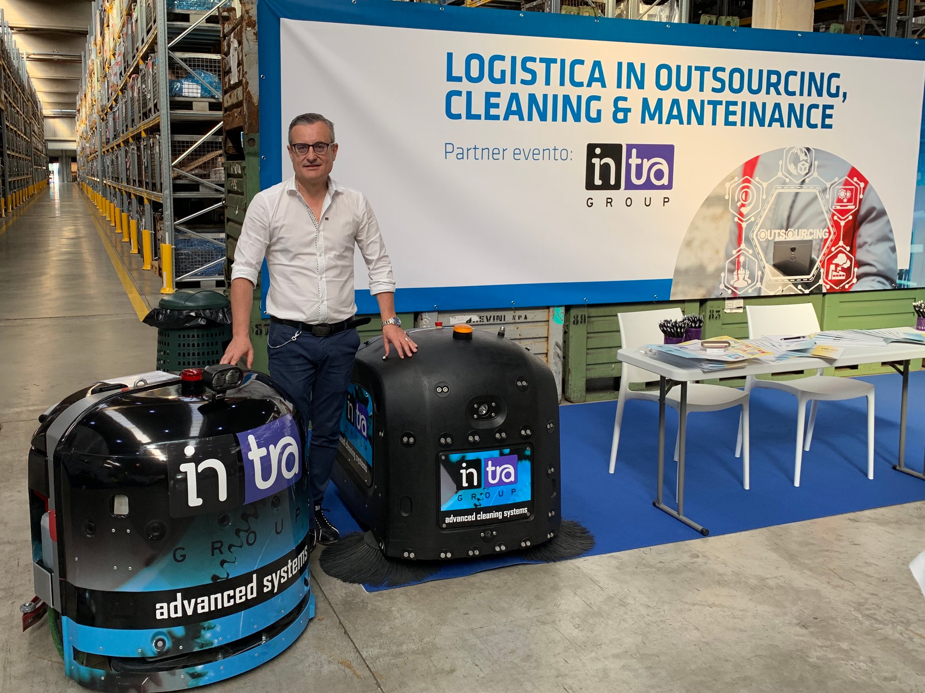 Un uomo posa con due robot aspirapolvere davanti a uno stand pubblicitario in un magazzino.