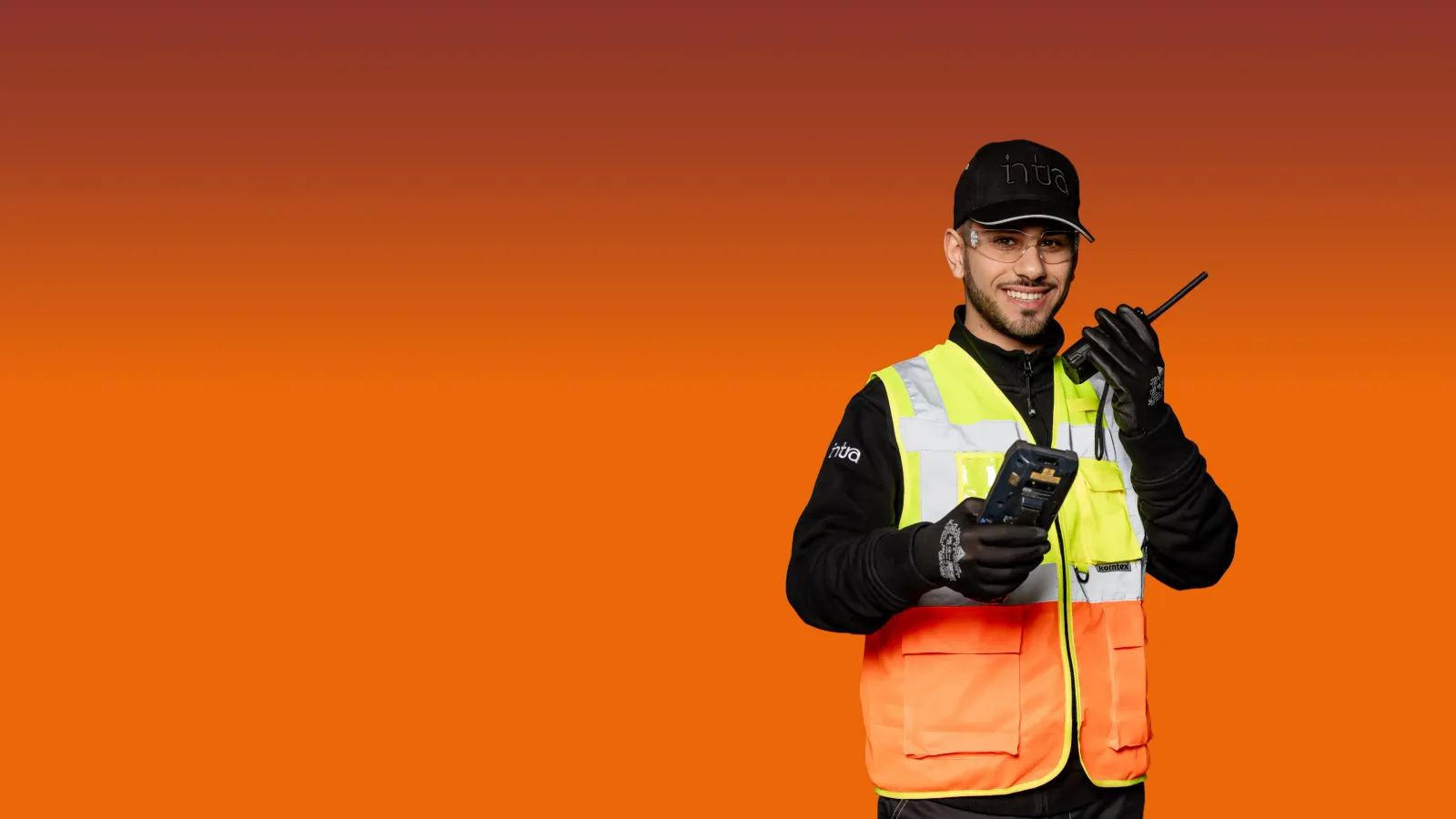 logistic_header.webp Un uomo sorridente con abbigliamento ad alta visibilità tiene in mano una radio e un dispositivo.