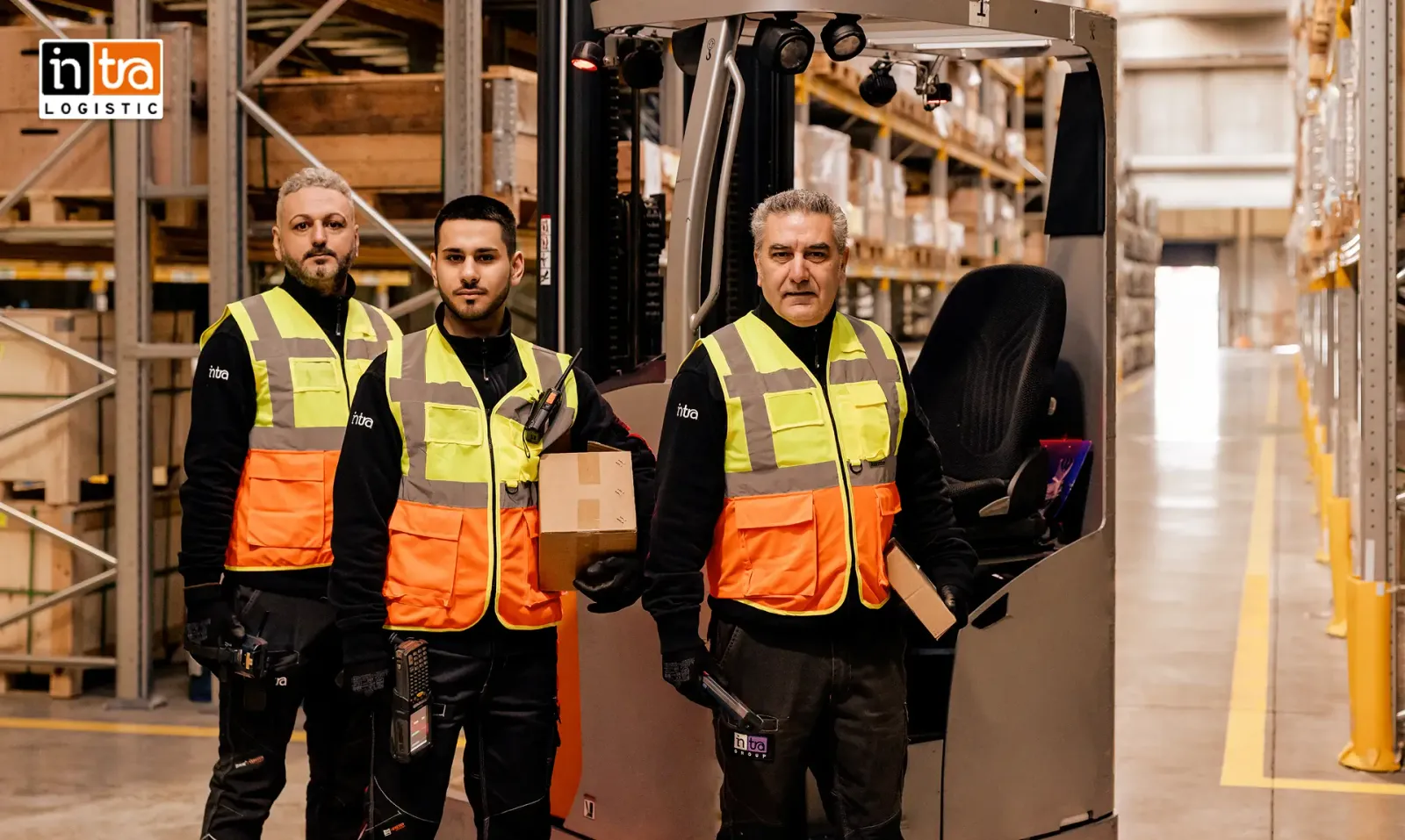 Ecco una possibile didascalia per l'immagine:

Tre dipendenti di Intra Logistics in un magazzino, che indossano abbigliamento protettivo.