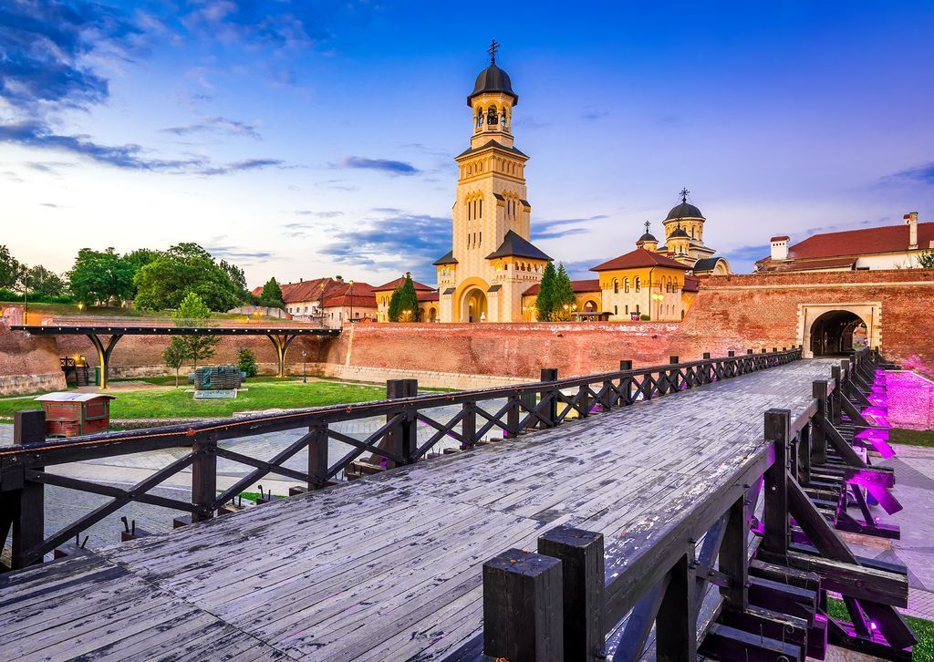 A wooden footbridge leading toward the stone walls and tall bell tower of the Alba Iulia Citadel at sunset.