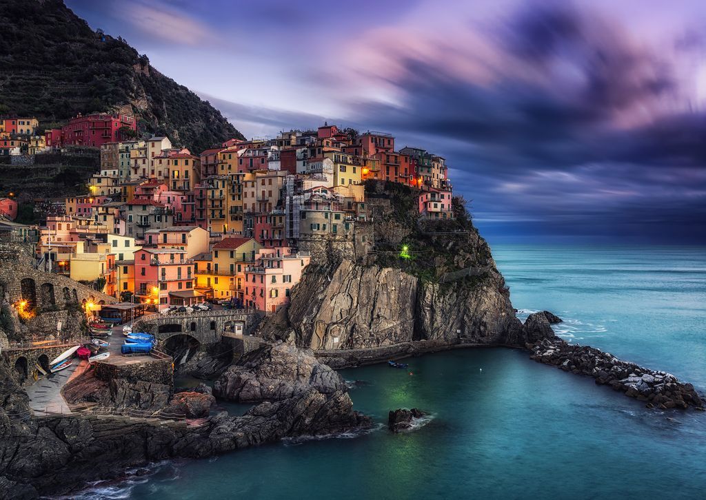 A colorful Italian coastal village perched on rocky cliffs above a turquoise sea at sunset.