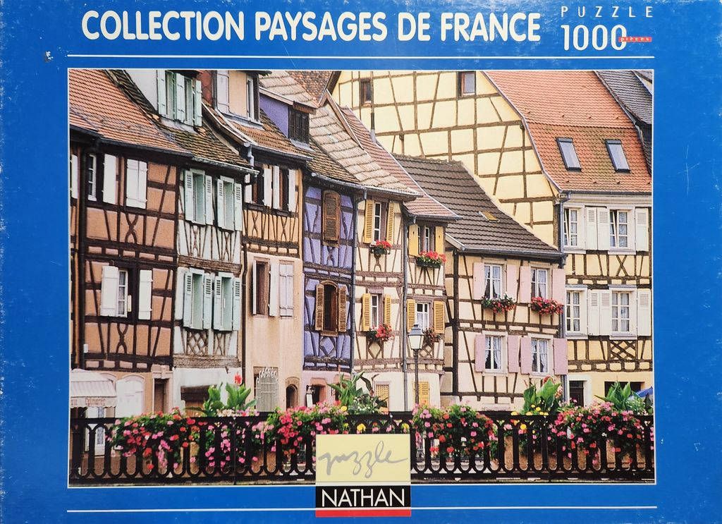 A row of colorful half-timbered houses in France with flower-lined balconies and a black iron railing in the foreground.