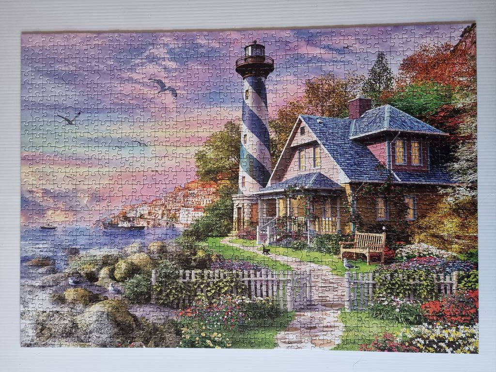 A lighthouse with a blue and white striped pattern stands beside a cozy cottage overlooking a sunset harbor.