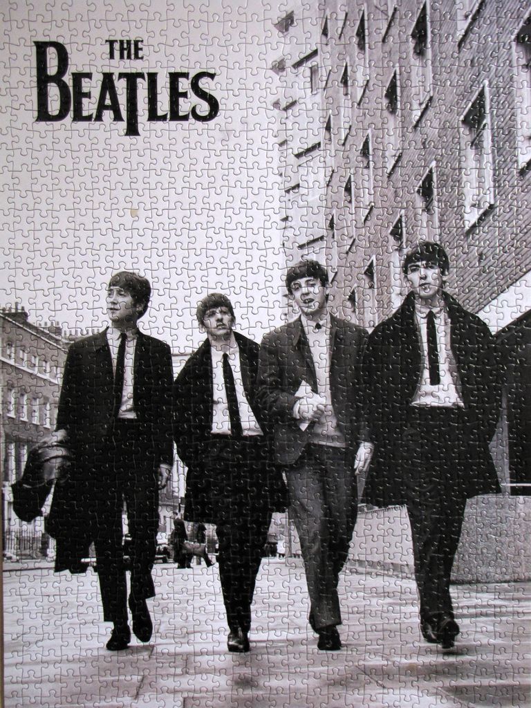 Black and white portrait of The Beatles walking down a city street in suits and long coats.